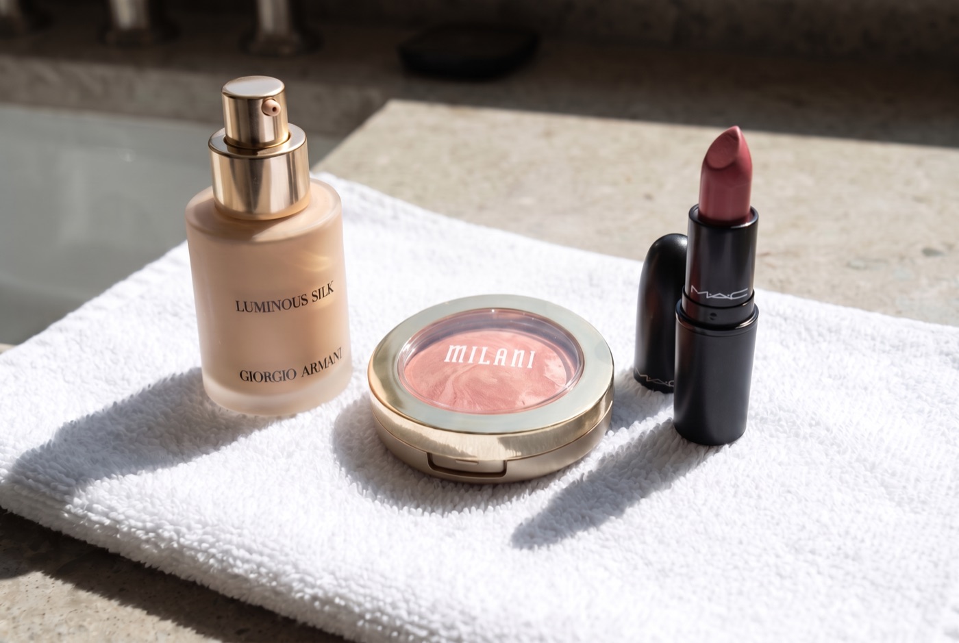 Giorgio Armani Luminous Silk Foundation, a Milani Baked Blush compact and a MAC lipstick on a white towel on a hotel vanity