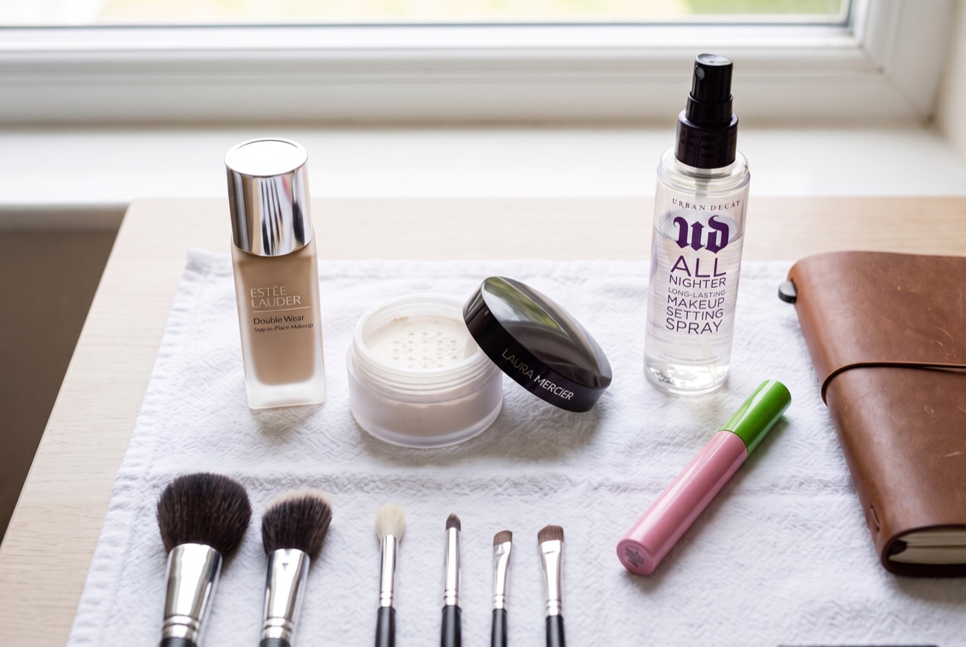 A bridal trial kit laid out on a white towel: Estée Lauder Double Wear, Laura Mercier setting powder, Urban Decay All Nighter setting spray, a waterproof mascara and a row of brushes