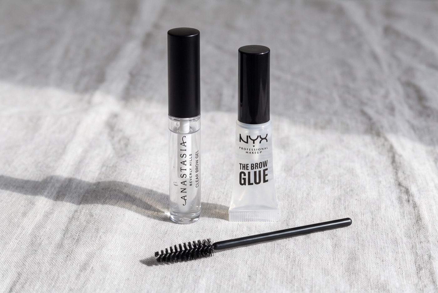 Anastasia Beverly Hills Clear Brow Gel and NYX The Brow Glue next to a black spoolie on a grey linen cloth
