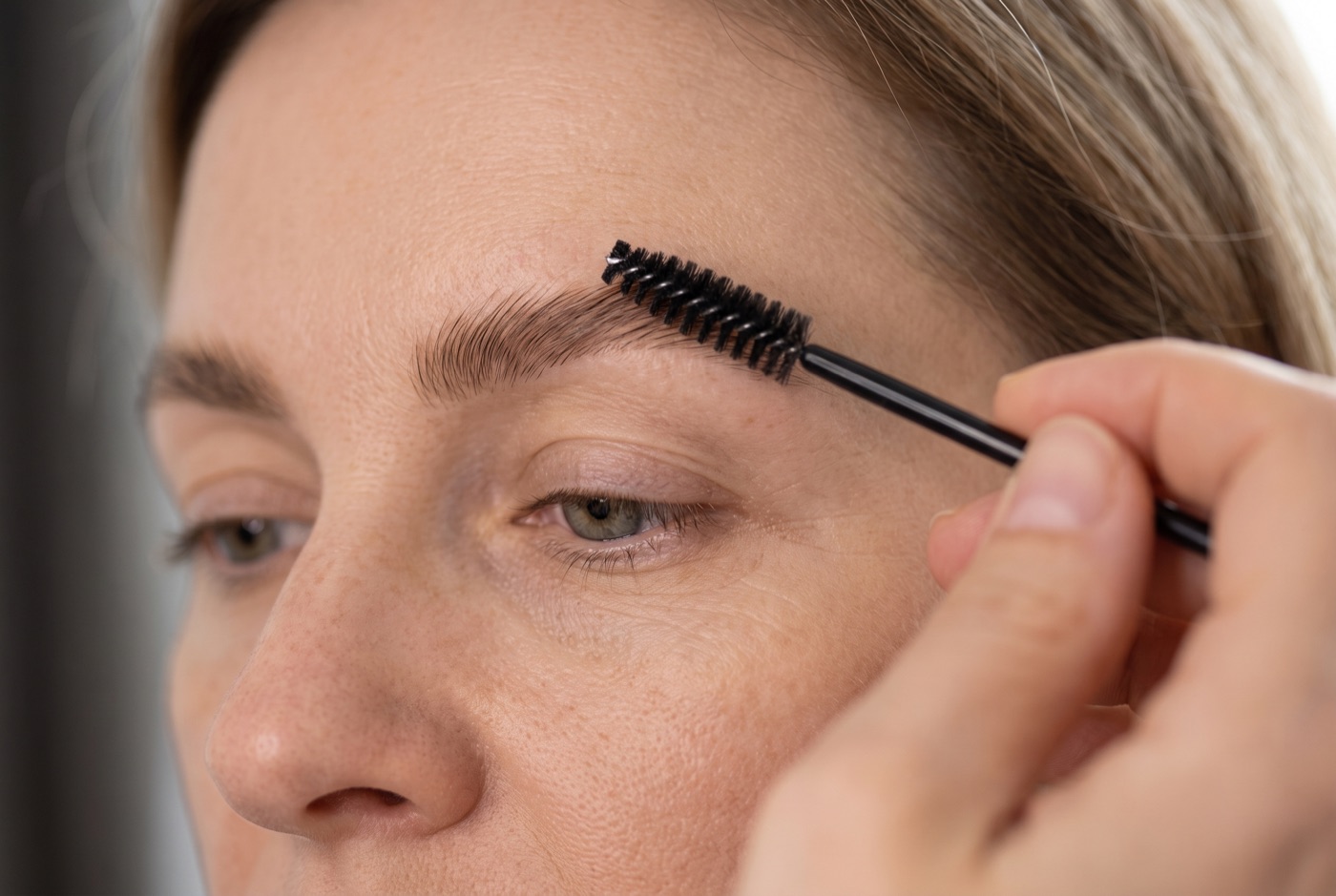 A makeup artist brushing a woman's natural brows upward with a clean black spoolie in soft window light