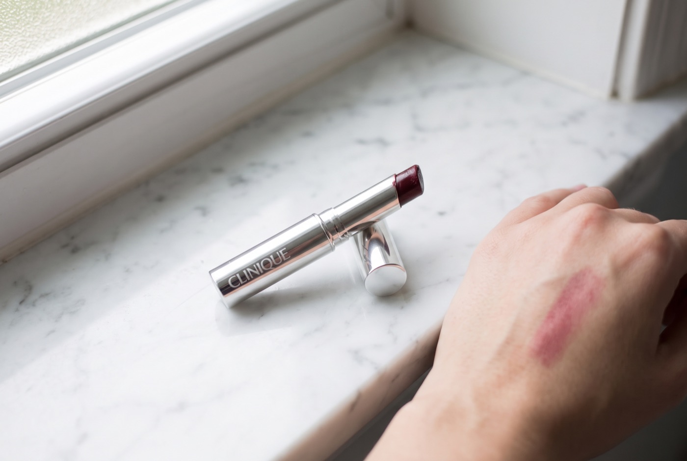 A silver Clinique Almost Lipstick in Black Honey with the cap off on a marble counter, next to a sheer berry swatch on the back of a hand