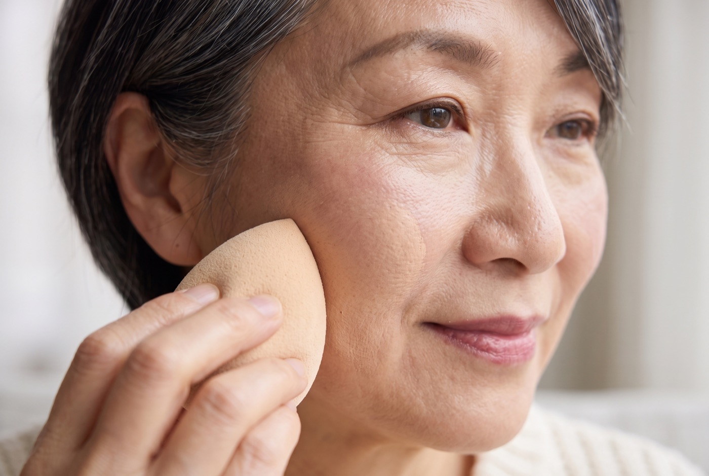 A damp beige makeup sponge pressing a sheer layer of foundation onto a woman's cheek, skin texture still visible
