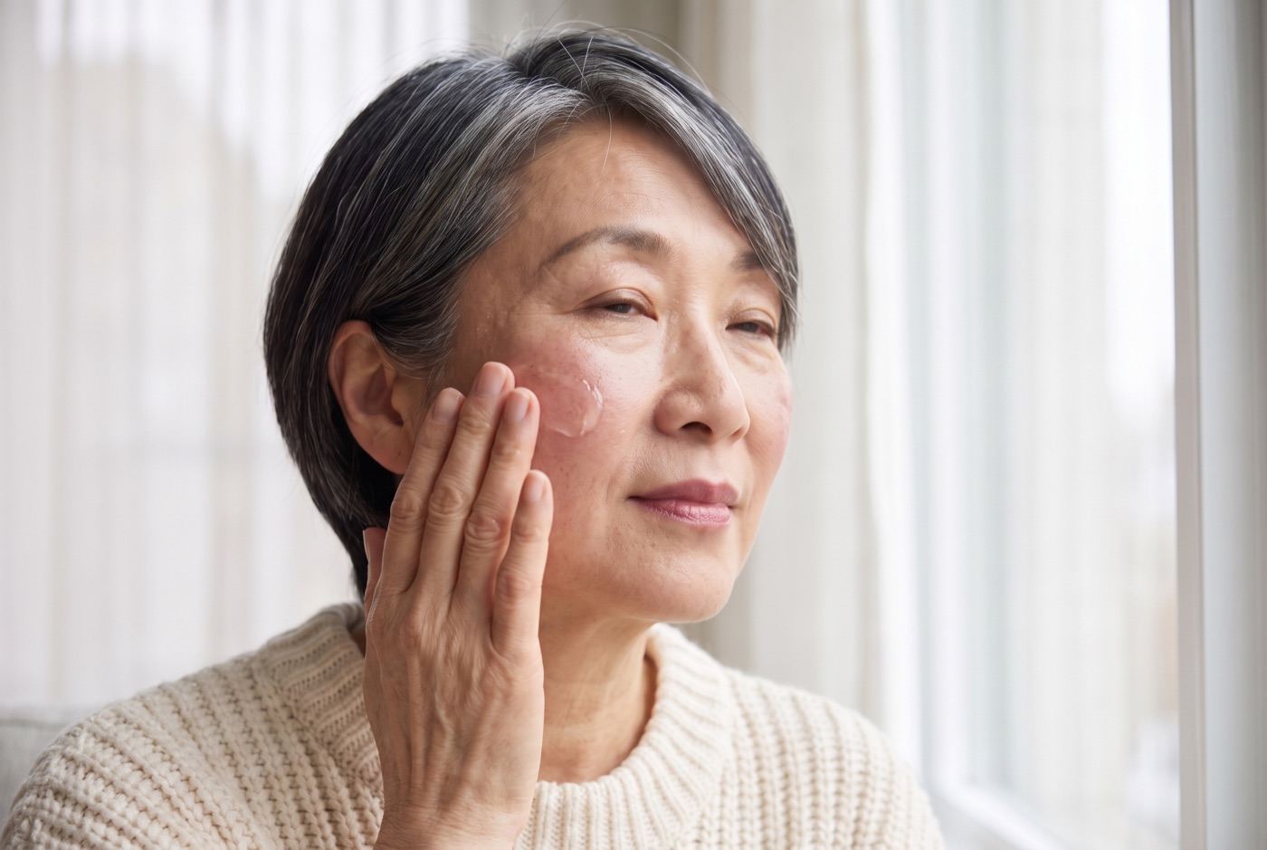 A woman in her sixties pressing a light gel moisturizer into her bare cheek with her fingertips by a window