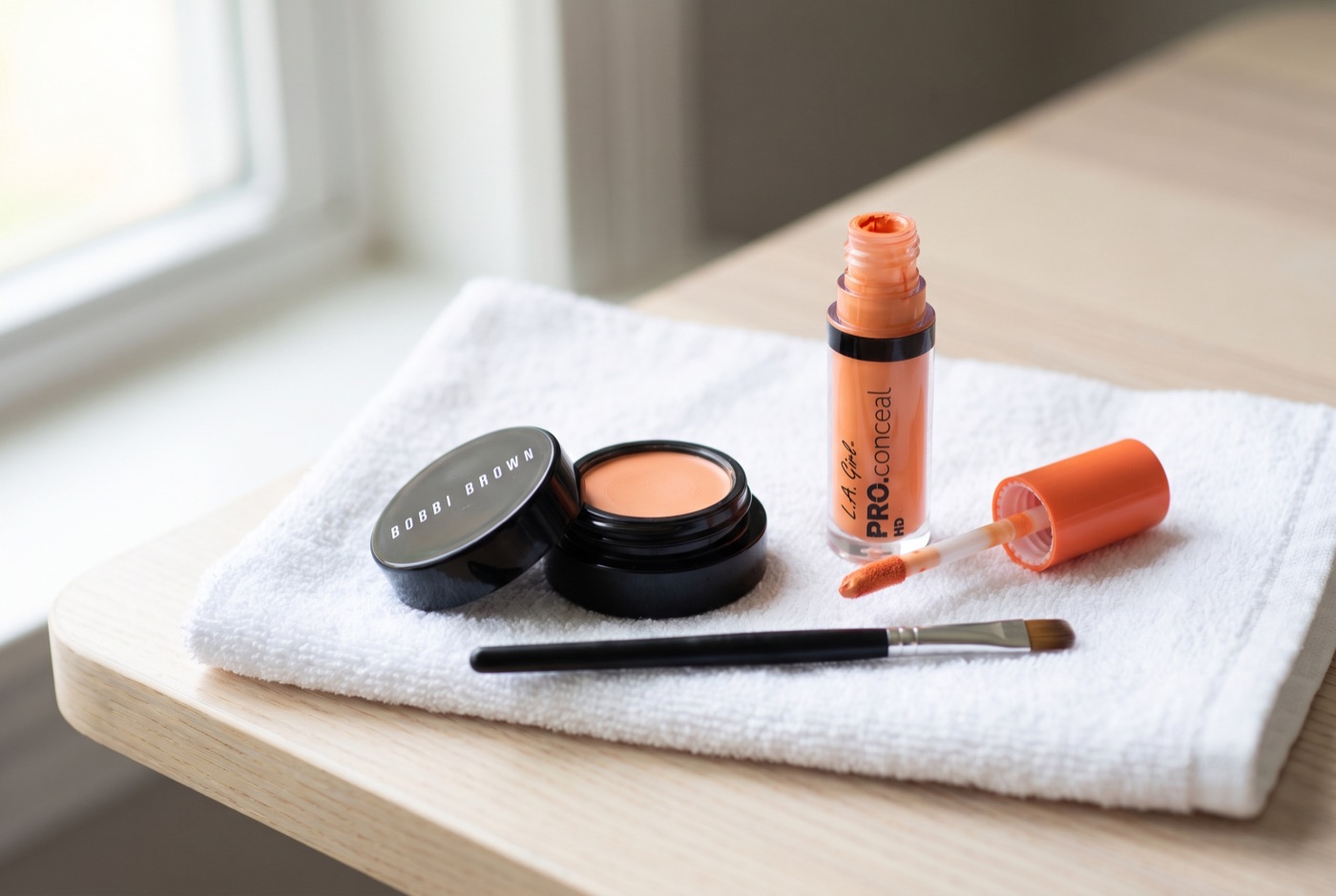 A small black pot of Bobbi Brown Corrector in peach and a bottle of L.A. Girl Pro Conceal HD orange corrector on a white towel