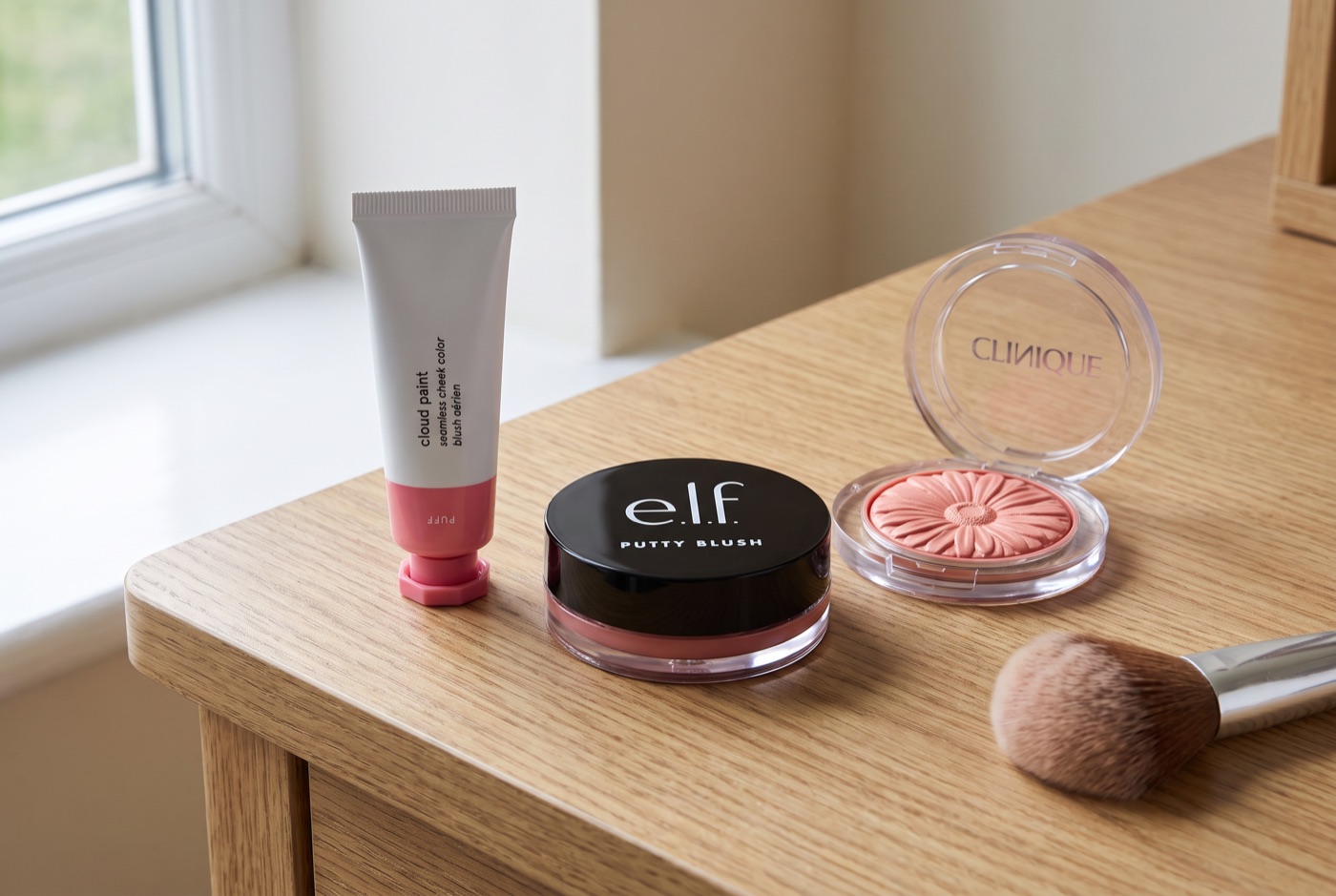 Glossier Cloud Paint, e.l.f. Putty Blush and Clinique Cheek Pop on a light oak dressing table with a fluffy blush brush