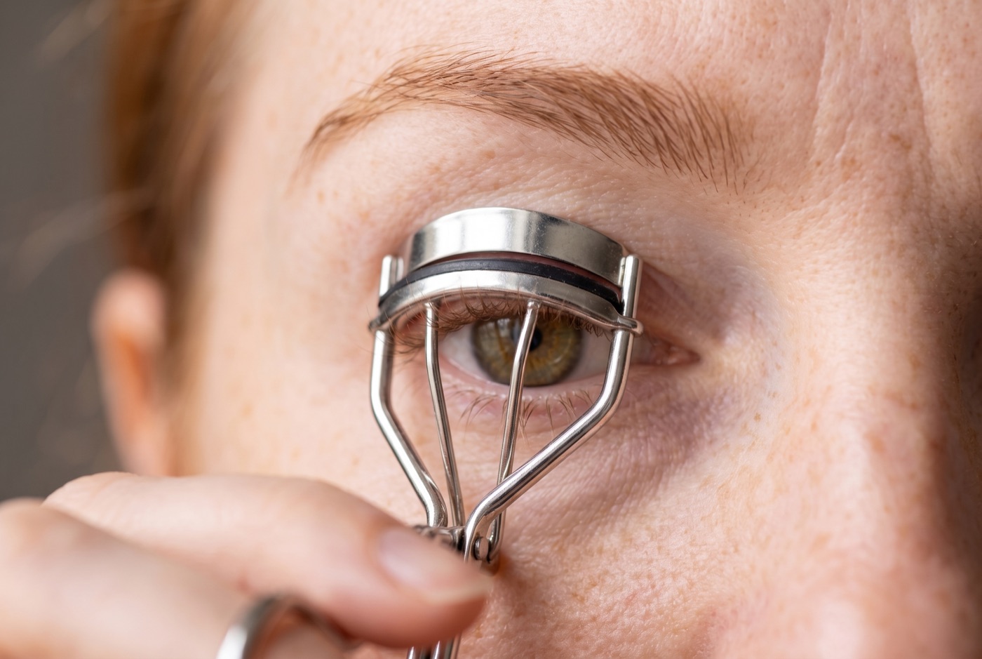 A plain steel eyelash curler clamped gently at the root of the upper lashes while the woman looks down