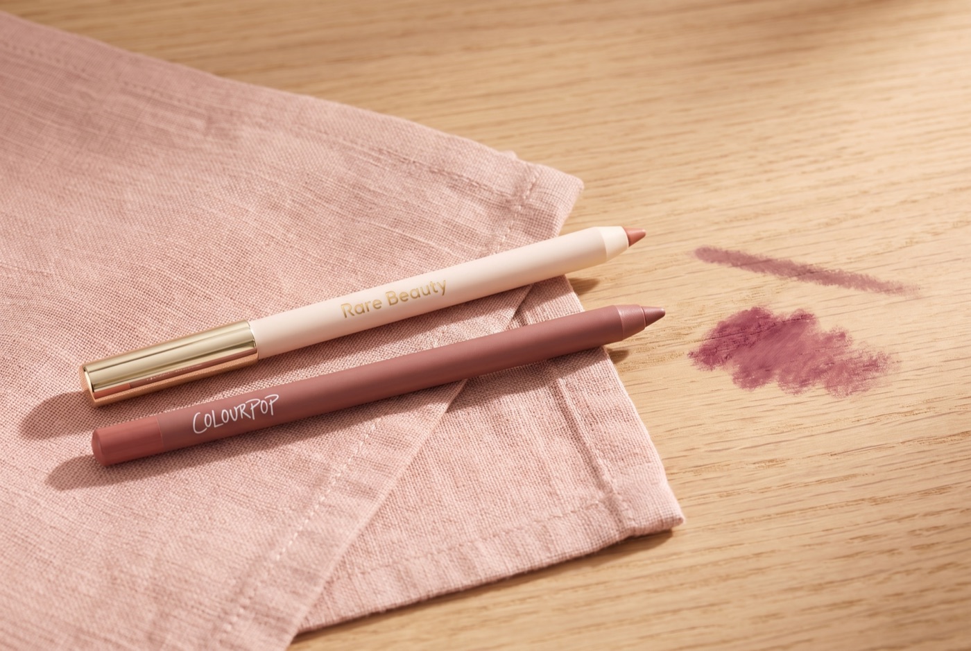 A Rare Beauty Kind Words matte lip liner and a ColourPop Lippie Pencil on a pink linen napkin with two soft blurred swatches