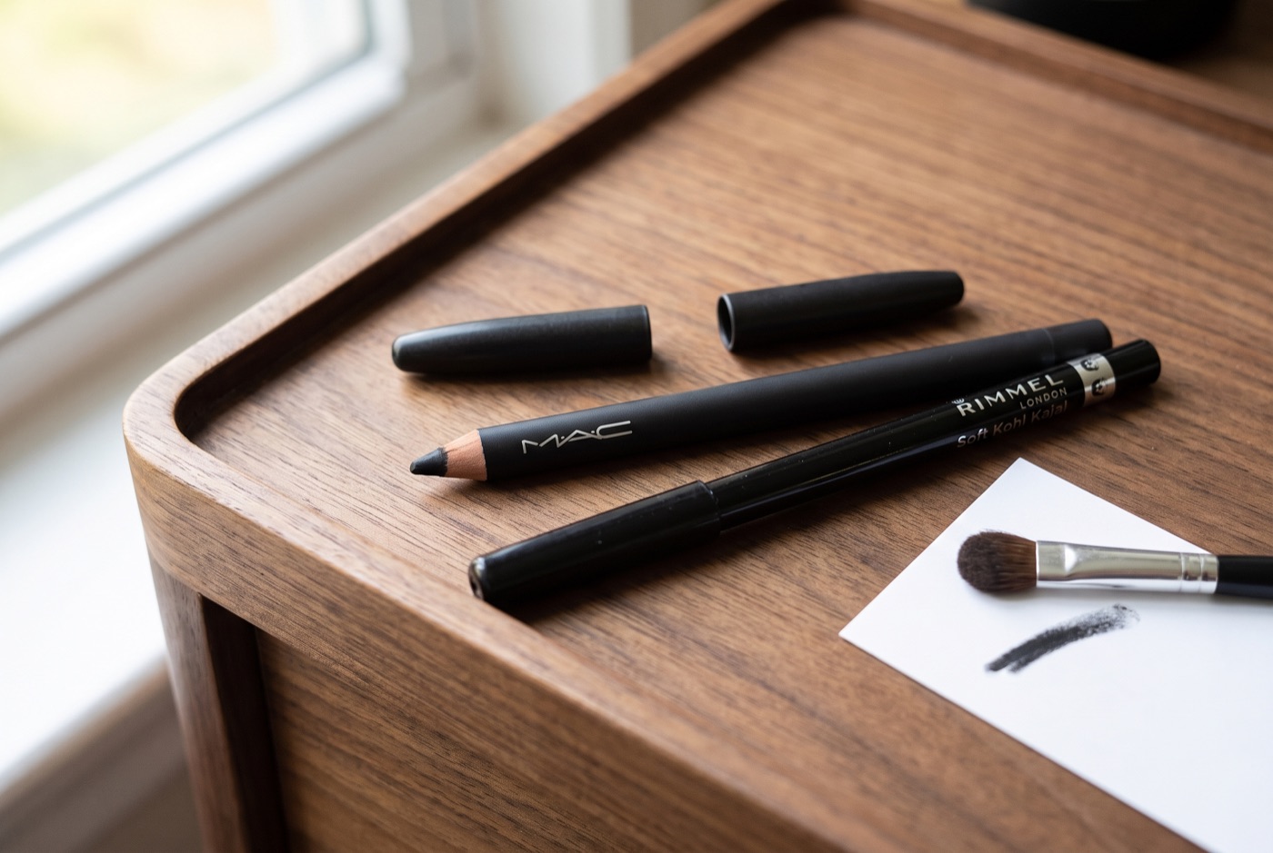 A black MAC Eye Kohl pencil and a Rimmel London Soft Kohl Kajal pencil on a walnut tray next to a small smudge brush