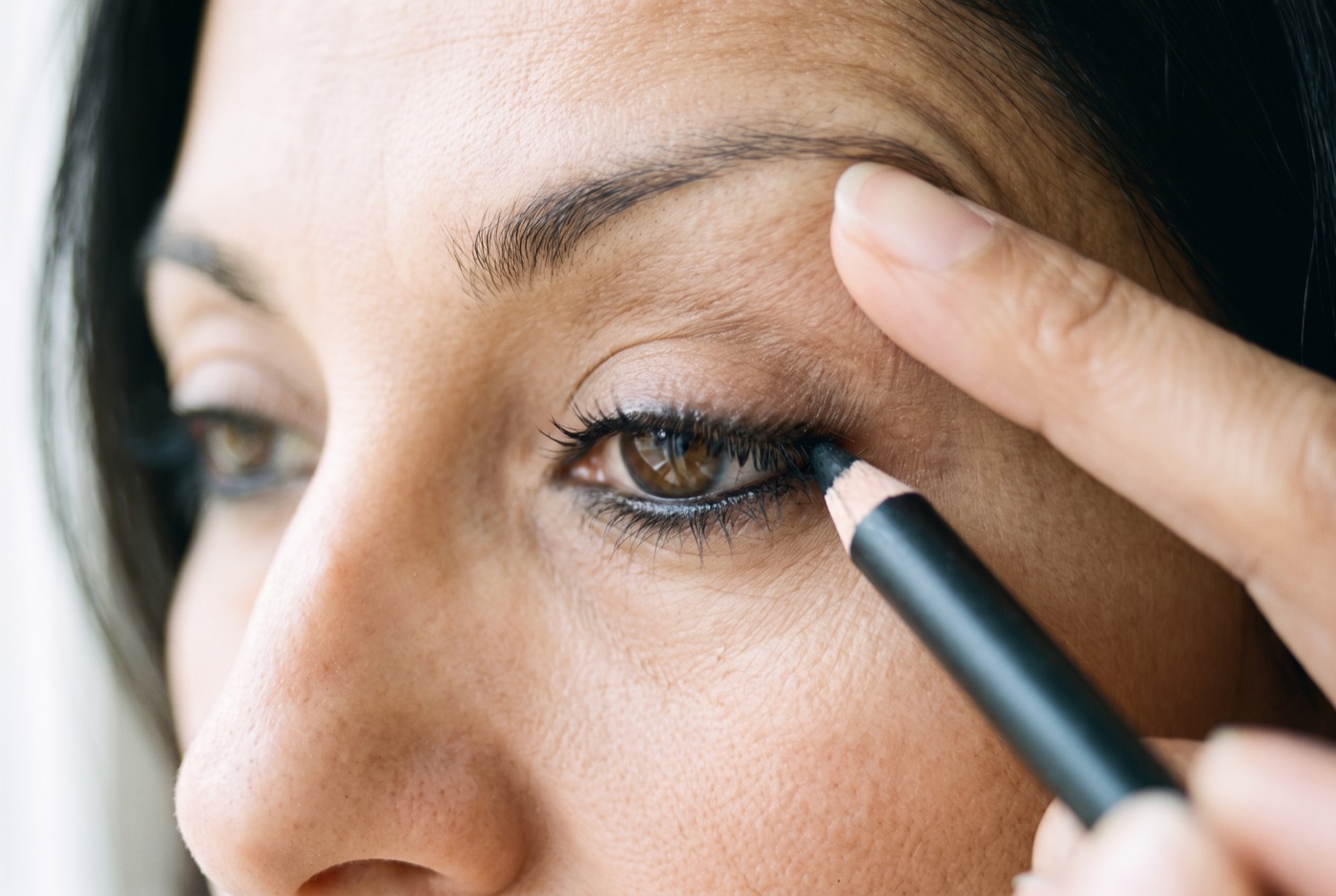 A fingertip lifting the upper lid while a black kohl pencil presses into the roots of the upper lashes at the outer corner