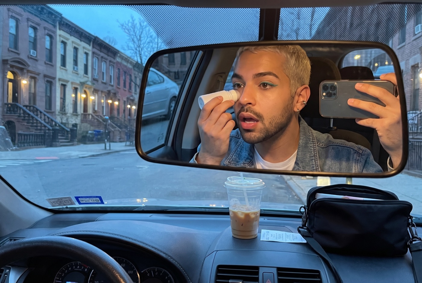 Danny in the driver's seat at dawn, seen in the rear-view mirror, rolling a metal eye roller under one eye, phone held up in the other hand