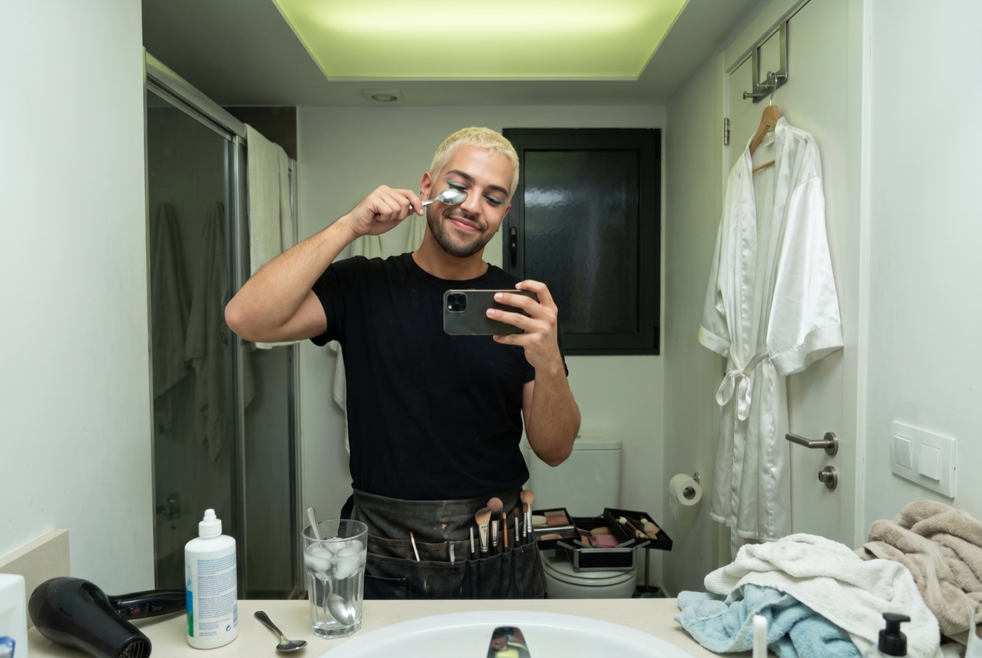 Danny Rose in a client's bathroom mirror before dawn, pressing the back of a cold metal spoon under one eye, phone in the other hand
