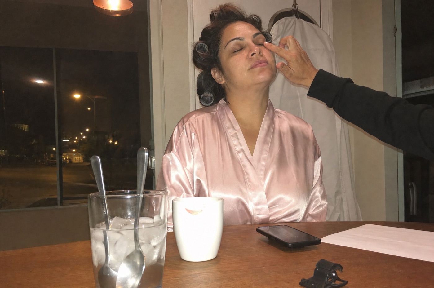A hand tapping with the ring finger under a bride's eye at a dining table before dawn, a glass of ice water with two spoons in it next to a coffee cup