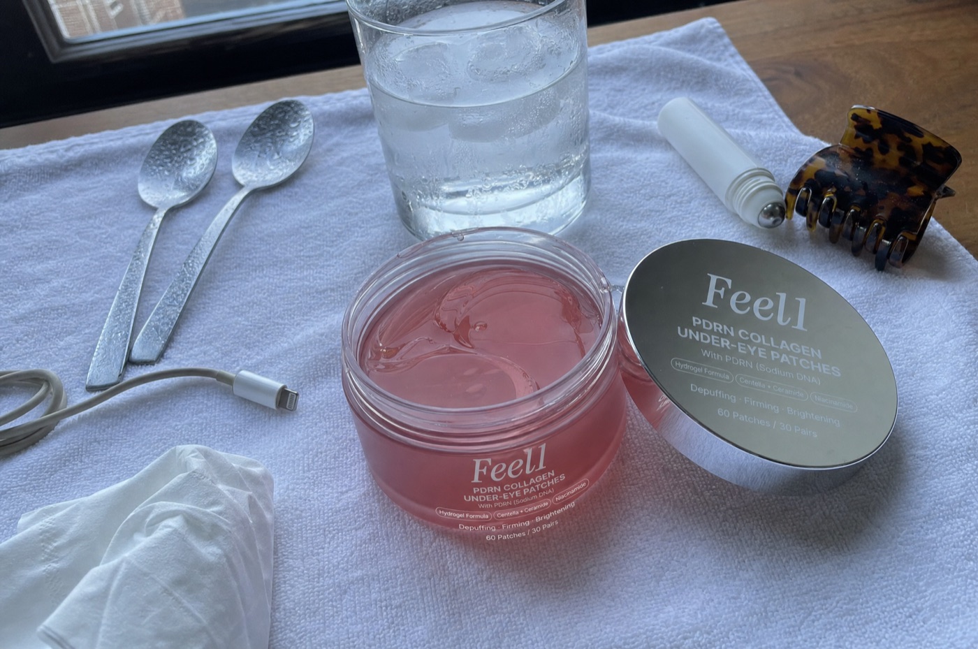 Two teaspoons, a caffeine eye roller with its label turned away, and an open jar of Feel1 PDRN patches on a white towel on a bride's vanity in morning window light
