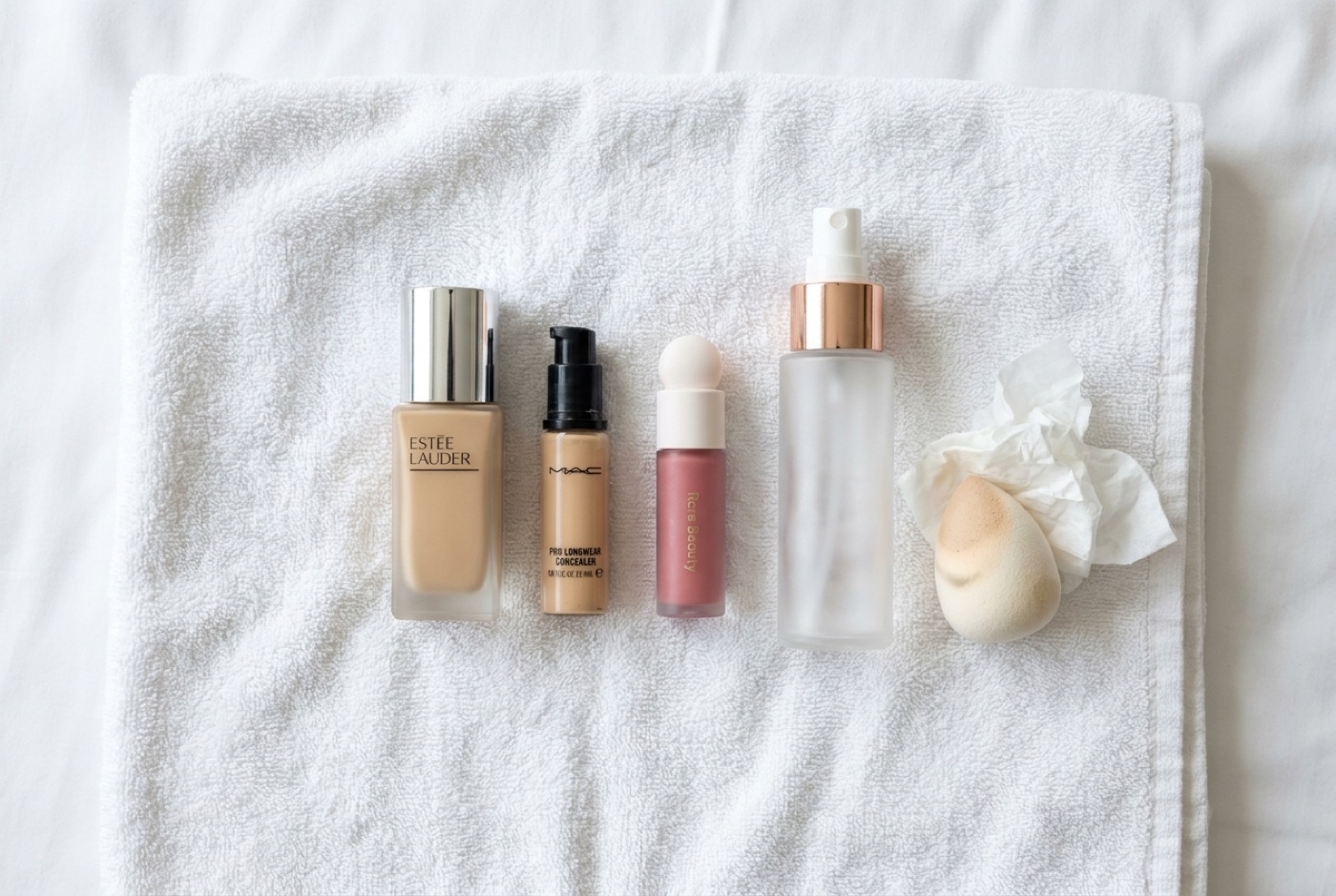 Estée Lauder Double Wear, MAC Pro Longwear Concealer, Rare Beauty Soft Pinch blush and Charlotte Tilbury setting spray laid out on a white towel