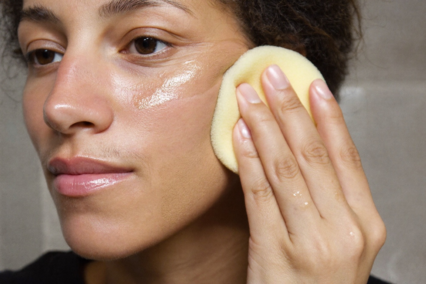 A woman patting foundation into her cheek with a damp sponge, half her face still bare