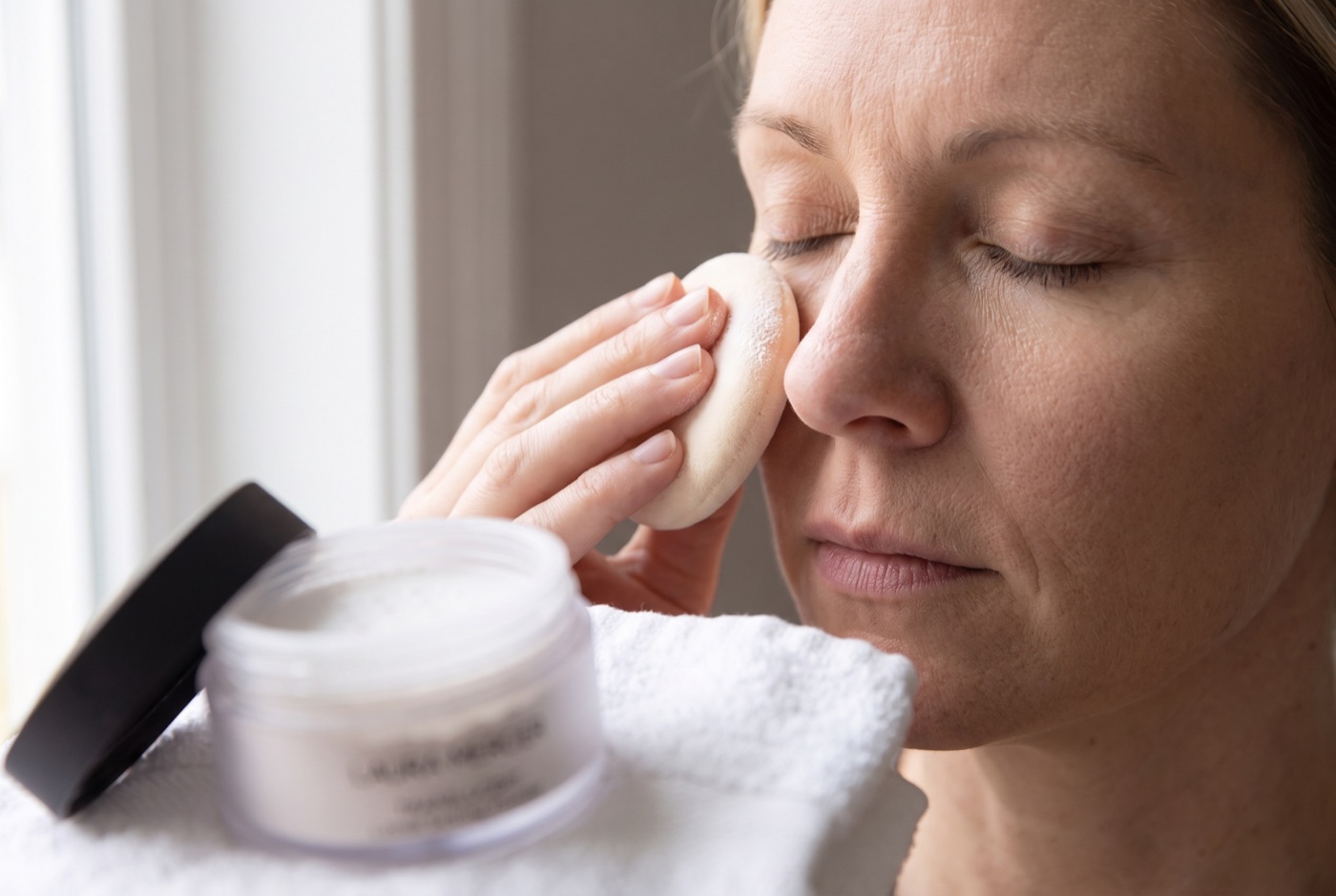 A velour puff pressed against the under-eye and the side of the nose, with the open jar of Laura Mercier setting powder in the foreground