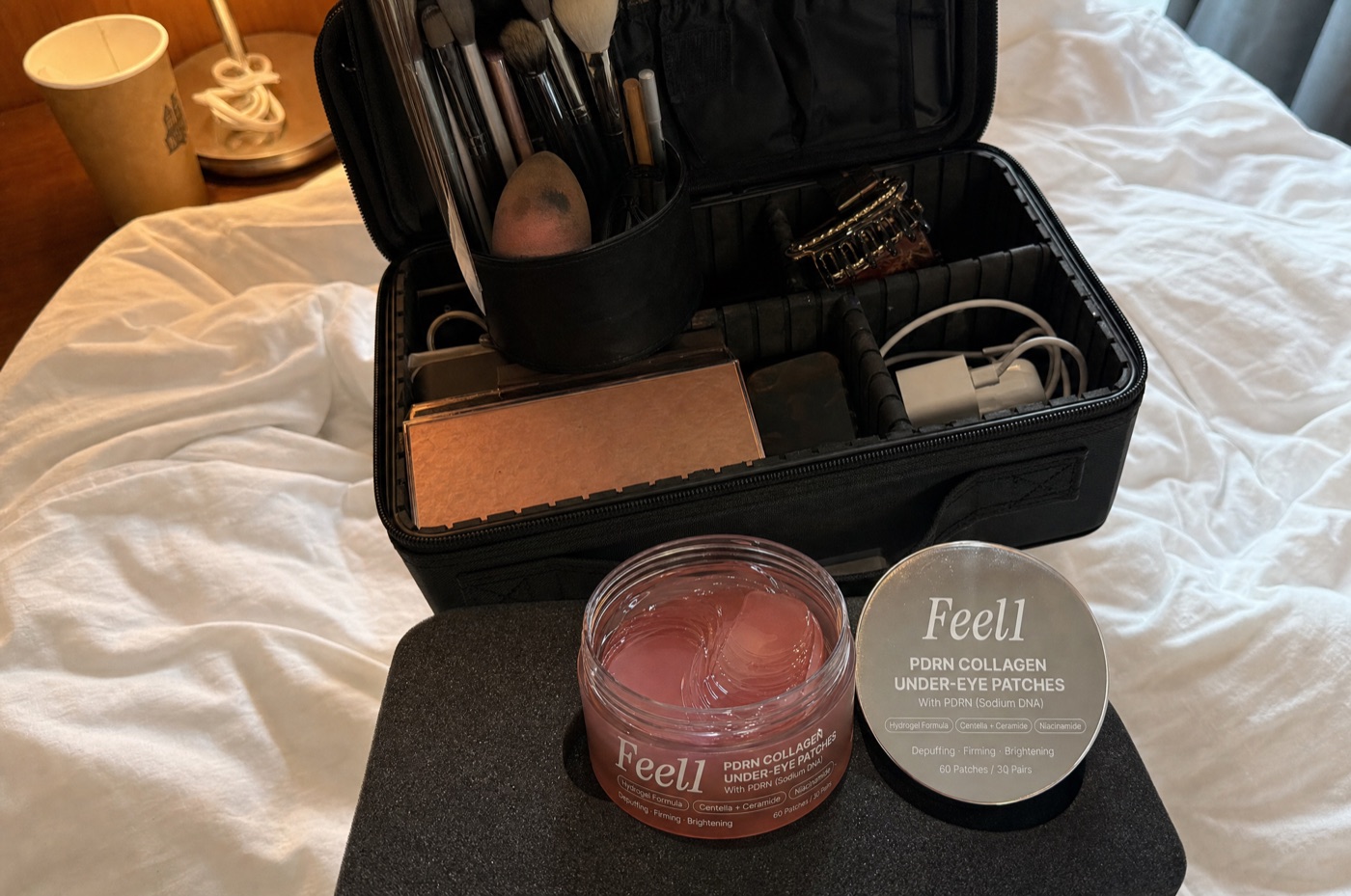 An open jar of Feel1 PDRN Collagen Under-Eye Patches with a chrome lid, pink hydrogel patches visible, inside an open makeup kit case on a hotel bed