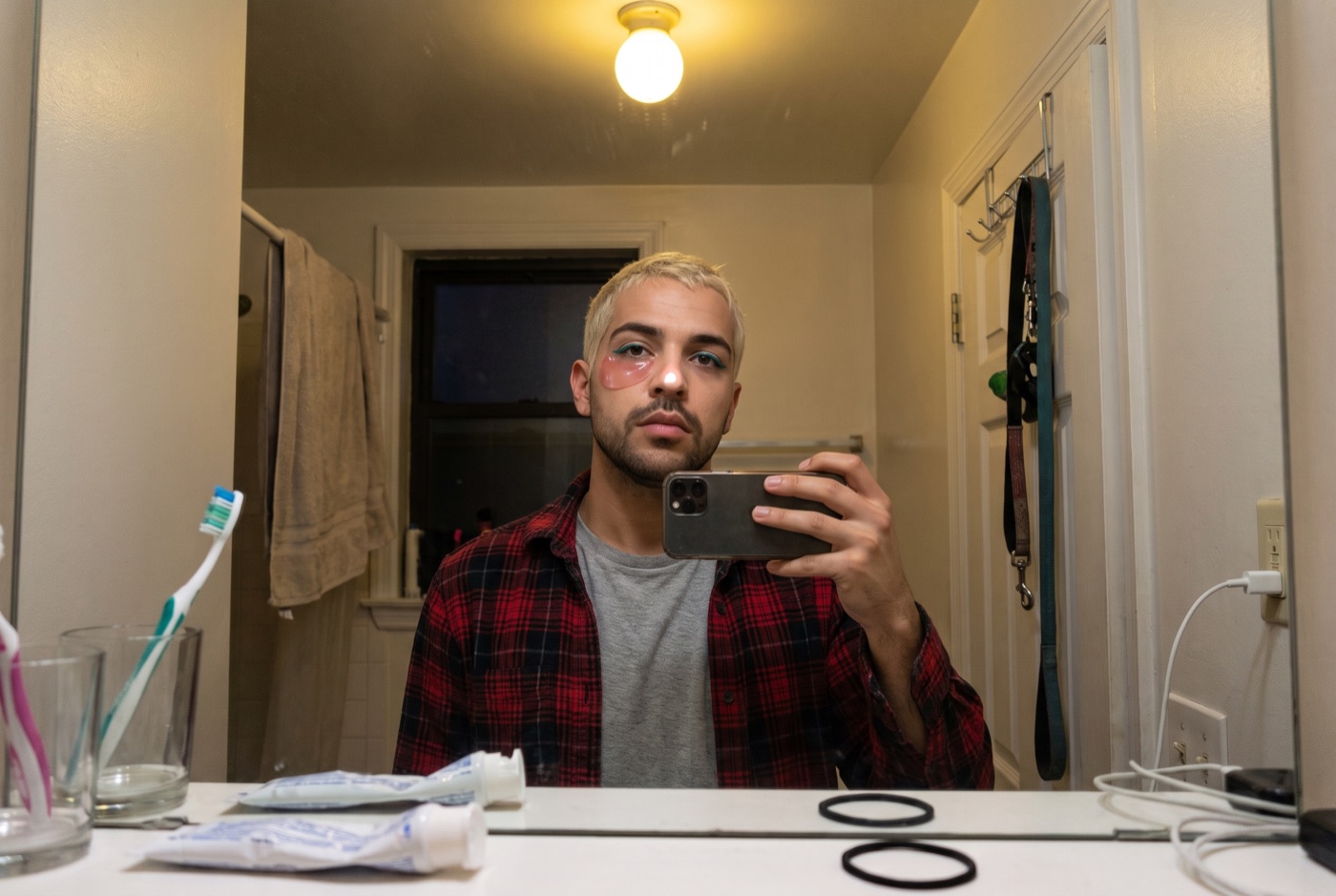 Danny in a bathroom mirror at six in the morning with a pink hydrogel patch under one eye and the other eye bare, phone in hand