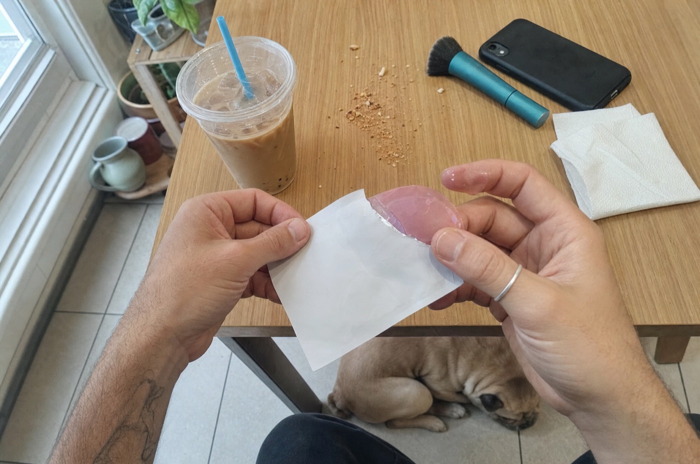 Hands tearing open a plain foil eye-patch sachet at a kitchen table in window light, gel on two fingers, a French bulldog asleep under the table