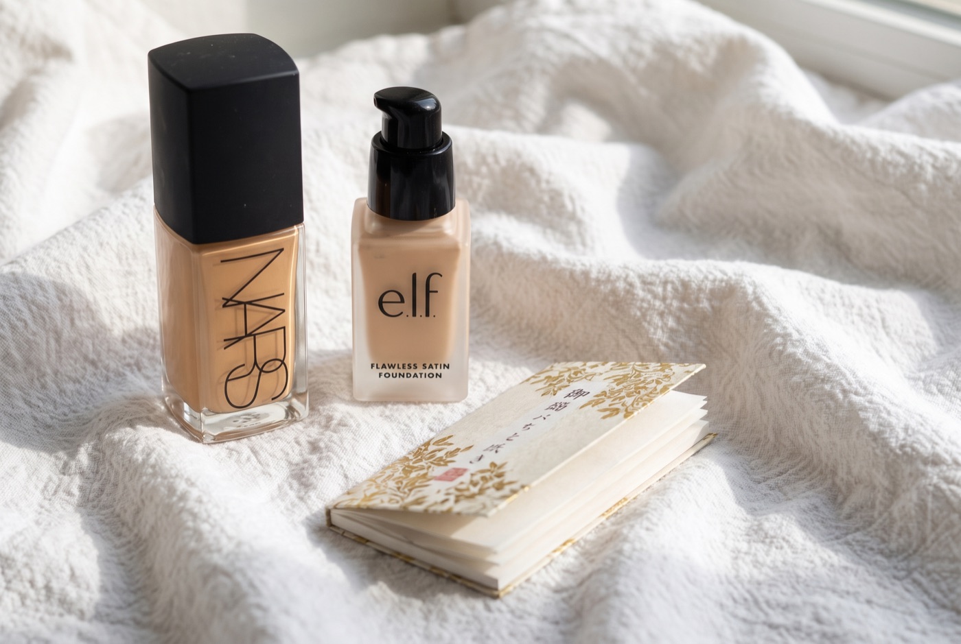Two foundation bottles, NARS Light Reflecting Foundation and e.l.f. Flawless Satin Foundation, beside a booklet of blotting papers on a white towel